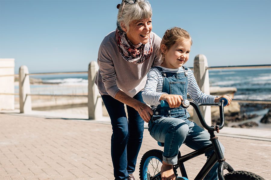 About Our Agency - Portrait of a Cheerful Grandmother Teaching Her Smiling Granddaughter How to Ride a Bike on the Boardwalk